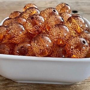 May include: A white bowl filled with amber-colored glass beads. The beads have a speckled, textured surface and are approximately 1 inch in diameter.