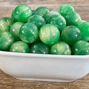 May include: A collection of green, marbled beads in a white bowl. The beads have a swirl pattern and varying shades of green. The bowl is white and has a simple, square shape. The beads are likely used for jewelry making or crafts.