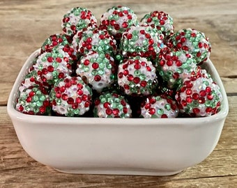 May include: A white ceramic bowl filled with decorative beads. The beads are white, covered in red and green rhinestones. The beads are round and have a textured surface. The bowl is square with rounded corners and sits on a wooden surface.