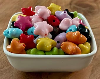 Può includere: Una ciotola quadrata bianca piena di perline colorate a forma di stella e cuore. Le perline sono di vari colori, tra cui rosso, rosa, giallo, arancione, blu, verde, viola e nero. La ciotola è appoggiata su una superficie di legno.
