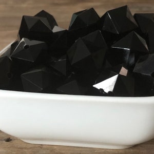 May include: A white ceramic bowl filled with black faceted beads. The beads are all the same size and shape, and they are arranged in a random pattern.