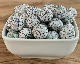 May include: A white ceramic bowl filled with iridescent, round beads. Each bead features a black spiderweb design. The beads have a pearlescent sheen, creating a shimmering effect. The bowl sits on a wooden surface.