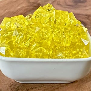 May include: A white square bowl filled with bright yellow, faceted, decorative glass crystals. The crystals have a sparkling, gem-like appearance. The bowl is sitting on a wooden surface.