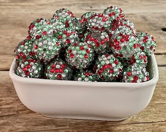 May include: A white ceramic bowl filled with a collection of large, round beads. The beads are covered in red, green, and silver rhinestones, creating a sparkling effect. The beads are likely for jewelry making or craft projects.