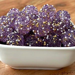 May include: A white square bowl filled with purple, spherical candies. The candies are covered in small white, gold, and silver embellishments. The bowl sits on a wooden surface.