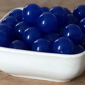 May include: A white square bowl filled with numerous glossy, deep blue beads. The beads are spherical and evenly sized, creating a visually appealing arrangement. The bowl sits on a wooden surface.