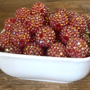 May include: A white square bowl filled with numerous red-colored beads. Each bead is covered in small, iridescent crystals that reflect light, creating a sparkling effect. The beads have a round shape and are clustered together.