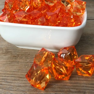 May include: A white bowl filled with orange faceted glass beads. The bowl is sitting on a wooden surface. Several orange faceted glass beads are scattered on the surface.