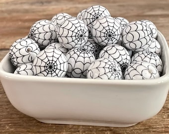 May include: A white ceramic bowl filled with round beads. Each bead is white with a black spiderweb design. The beads are a Halloween or spooky-themed craft supply.
