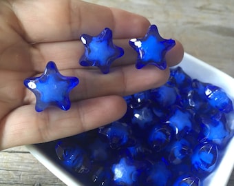 May include: A hand holds three translucent, star-shaped beads with a deep blue hue. A white square bowl is filled with similar beads. The beads have a faceted appearance, catching the light and creating a sparkling effect. These could be used for jewelry making or craft projects.