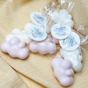 May include: Cloud-shaped soaps in pink and white, packaged in clear cellophane bags. Each soap has a round tag that reads "on cloud nine" and "Olivia's Bridal Shower 05.18.2025". The soaps are arranged on a beige fabric.