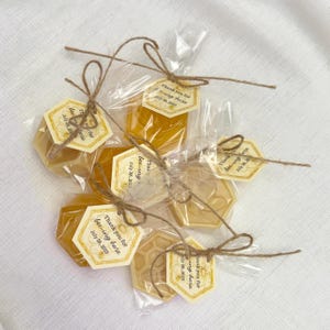 May include: Golden honey favors in hexagonal shapes, wrapped in cellophane and tied with twine. Each favor has a tag saying "Thank you for bee-ing here July 28, 2025". Some have a honeycomb design.