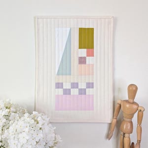 May include: A framed, quilted wall hanging with a geometric design in pastel colours. The artwork features a white background with sections of white, light blue, olive green, pink, and lavender. The piece is displayed on a white wall above a wooden shelf with a vase of white flowers and a wooden artist's mannequin.