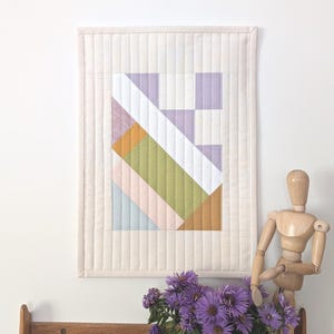 May include: A pastel-coloured geometric quilt hanging on a wall. The artwork features lavender, green, and peach hues. Below, a wooden shelf displays a terracotta sun sculpture, a vase of purple flowers, and a wooden artist's mannequin.