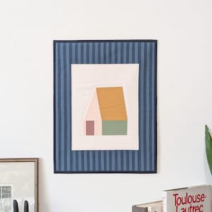 Peut inclure: Une tenture murale rectangulaire en tissu présentant un panneau central crème avec un motif de maison stylisé dans des tons rose, jaune moutarde et vert sauge. Le panneau central est encadré par une bordure à rayures verticales bleu foncé et bleu clair.
