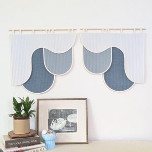 May include: Two abstract wall hangings with a layered design in shades of blue and white, mounted on wooden rods. The pieces have a quilted texture and a curved, modern shape. A framed print and potted plant are below.