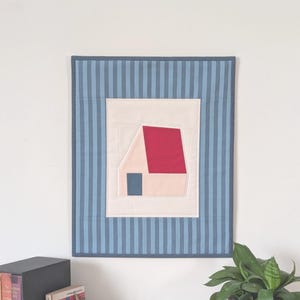 May include: A geometric house design wall hanging. The artwork features a red roof, a blue door, and a light pink wall, set against a white background. The border is blue with vertical stripes.