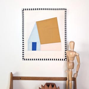 Peut inclure: Tableau abstrait géométrique avec un motif en forme de maison, comprenant un carré couleur moutarde et un toit bleu clair. L'œuvre est encadrée d'une bordure pointillée noire et blanche. Un mannequin d'artiste en bois et une étagère avec une pièce en terre cuite en forme de soleil sont également visibles.