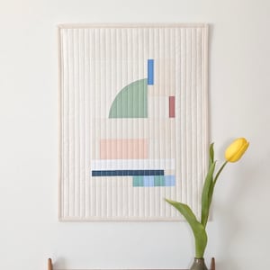 May include: A framed, quilted wall hanging with an abstract geometric design in pastel colours. The design includes shapes in green, pink, blue, and white. A yellow tulip in a vase sits below the artwork.