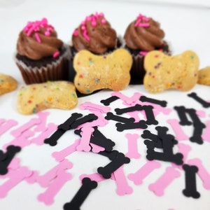 May include: Pink and black bone-shaped confetti scattered around chocolate cupcakes with pink sprinkles and bone-shaped cookies with colorful sprinkles.