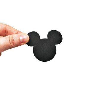 May include: A black, die-cut Mickey Mouse head shape, held against a white background. The iconic silhouette features two large circular ears atop a rounded head. The shape is made of a matte material.