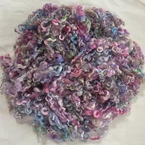 May include: A large pile of colorful, curly yarn in shades of pink, purple, blue, and green. The yarn has a soft, textured appearance, with a mix of light and dark tones. The yarn is likely used for knitting or crocheting projects.