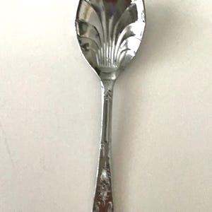 May include: A silver spoon with a decorative floral pattern on the handle and a fan-shaped design on the bowl. The spoon is made of a shiny, reflective metal, likely stainless steel.