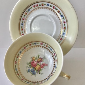 May include: A vintage teacup and saucer set. The cup and saucer are cream-colored with gold trim and a floral design in the center of the cup. The saucer has a matching floral and gold trim design.
