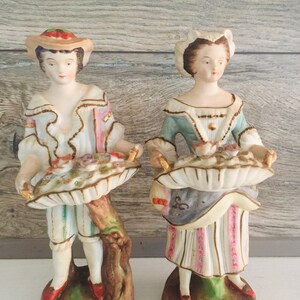 May include: A pair of vintage porcelain figurines of a man and woman in 18th-century clothing. They are both holding baskets filled with flowers. The man is wearing a blue and white striped jacket and pants, and the woman is wearing a blue and white dress with a pink and white striped skirt.