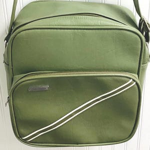 May include: A sage green shoulder bag with a rectangular silhouette, featuring a top zipper closure, a front zippered pocket, and two diagonal white stripes across the lower front. A small silver Samsonite logo plate is attached to the front.