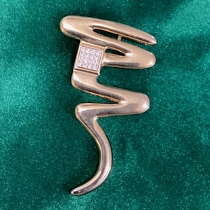 May include: A gold-toned brooch with a unique, abstract design. The brooch features a square section encrusted with small, clear stones, and a flowing, curved shape. The brooch is set against a dark green background.