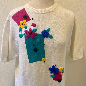 May include: Off-white short-sleeve sweater with colorful floral embroidery. The design features a teal square, a pink square, and a red pot, each adorned with yellow, pink, and blue flowers. The sweater is displayed on a mannequin.