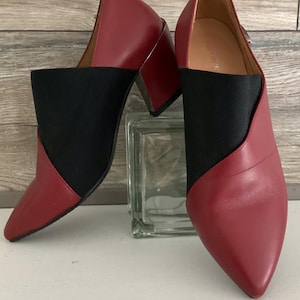 May include: A pair of pointed-toe shoes featuring a two-tone design with deep red leather panels and a contrasting black elasticized fabric center. The shoes have a sturdy block heel and a sleek, modern silhouette with a clean, minimalist aesthetic.