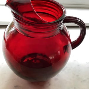 May include: A round, deep red glass pitcher featuring a wide, circular body, a prominent pouring spout, and a curved handle attached to the side. The glass is translucent, allowing light to pass through the vibrant red color of the vessel.