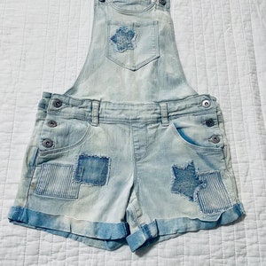 May include: Light blue denim overalls with a patchwork design. The overalls have a star-shaped patch on the front pocket and a cuffed hem.