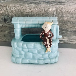 May include: A light blue ceramic planter shaped like a well with a small elf figurine sitting on the edge. The elf is wearing a brown robe and has a white face.