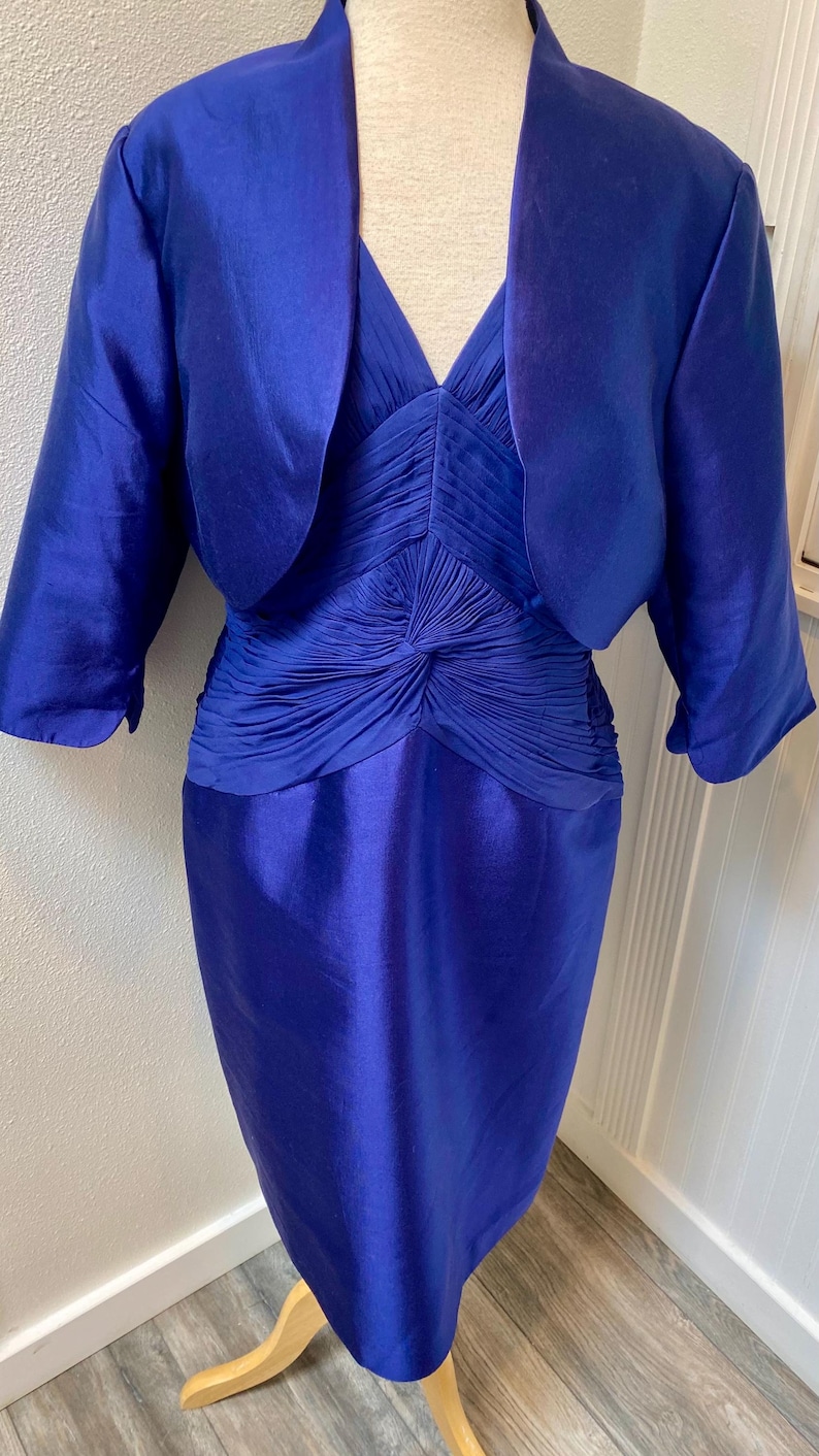 May include: A royal blue dress and matching bolero jacket. The dress has a fitted bodice with a gathered detail at the waist and a straight skirt. The bolero has a shawl collar and three-quarter sleeves.