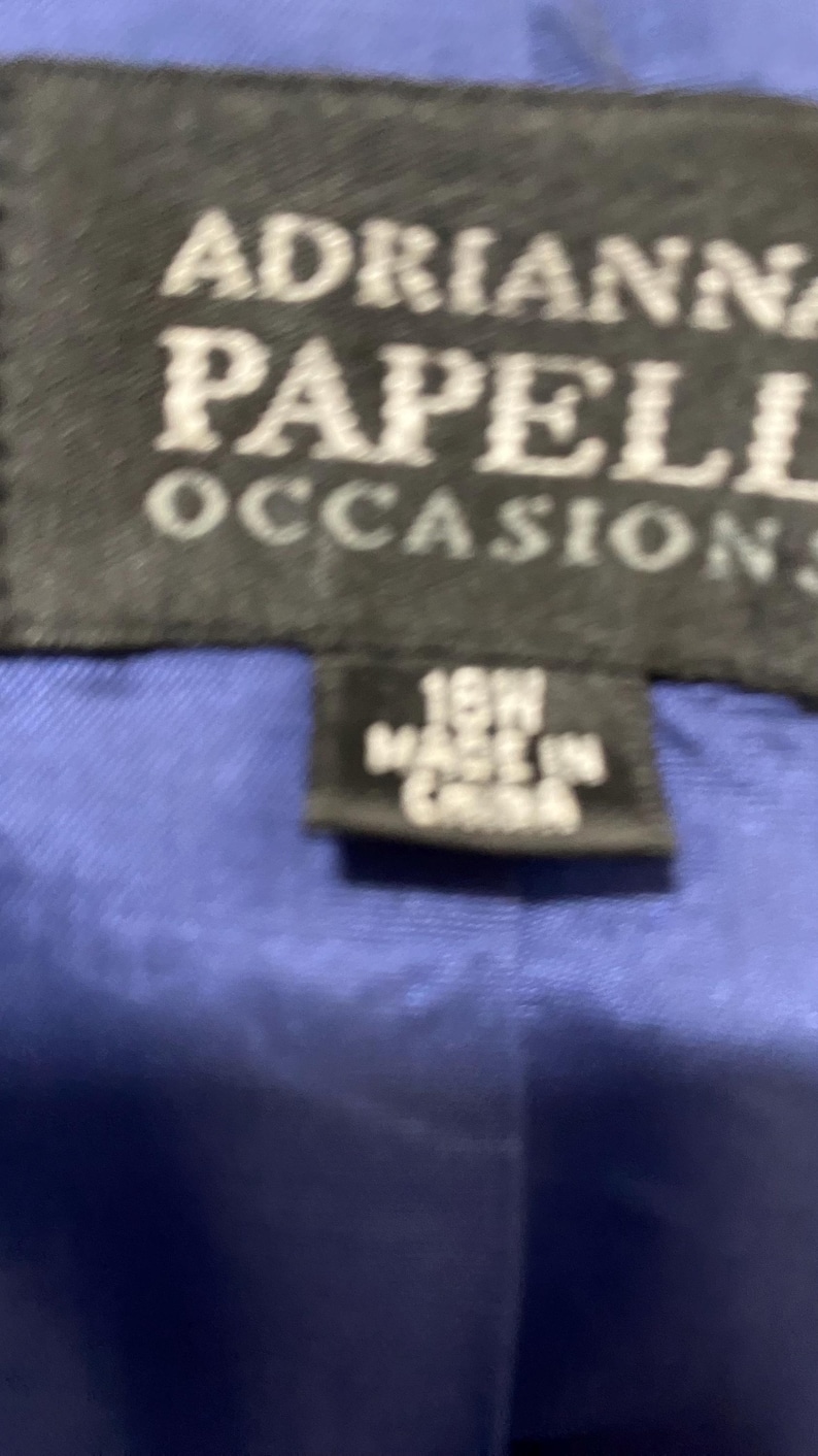 May include: A black label with white text that reads "Adrianna Papell Occasions" and a smaller label that reads "16W Made in China". The labels are on a blue fabric background.