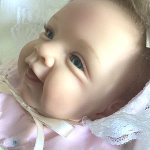 May include: A lifelike baby doll with blue eyes and a smiling expression. The doll is wearing a pink dress with a white lace collar and a matching bonnet. A white ribbon is tied around the neck.