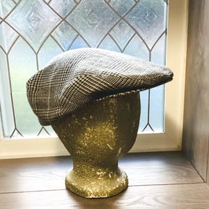 May include: A black and white plaid newsboy cap displayed on a vintage green mannequin head. The cap has a classic design and is made of a textured fabric. The background includes a window with a decorative pattern and wooden flooring.