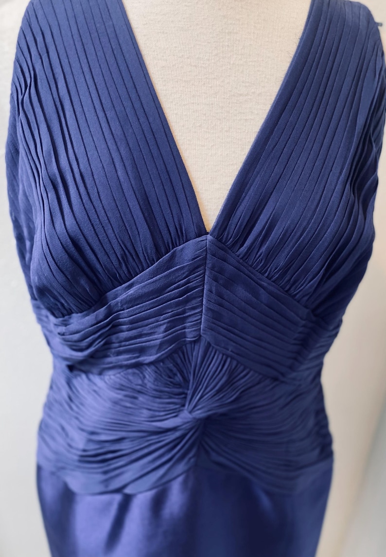 May include: A sleeveless, deep blue, pleated dress with a V-neckline. The dress is made of a silky fabric and has a fitted silhouette.