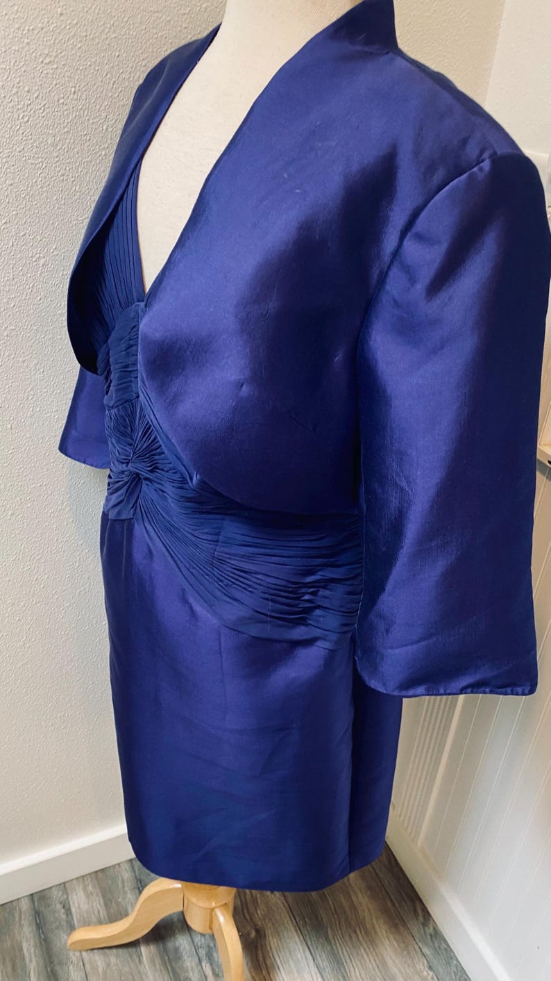 May include: A royal blue dress with a matching jacket. The dress has a fitted bodice with a pleated skirt and a V-neckline. The jacket has a shawl collar and three-quarter sleeves.