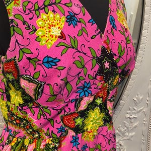 May include: A sleeveless pink dress with a V-neckline and a colorful floral print. The print features green, yellow, blue, red, and black flowers and leaves.