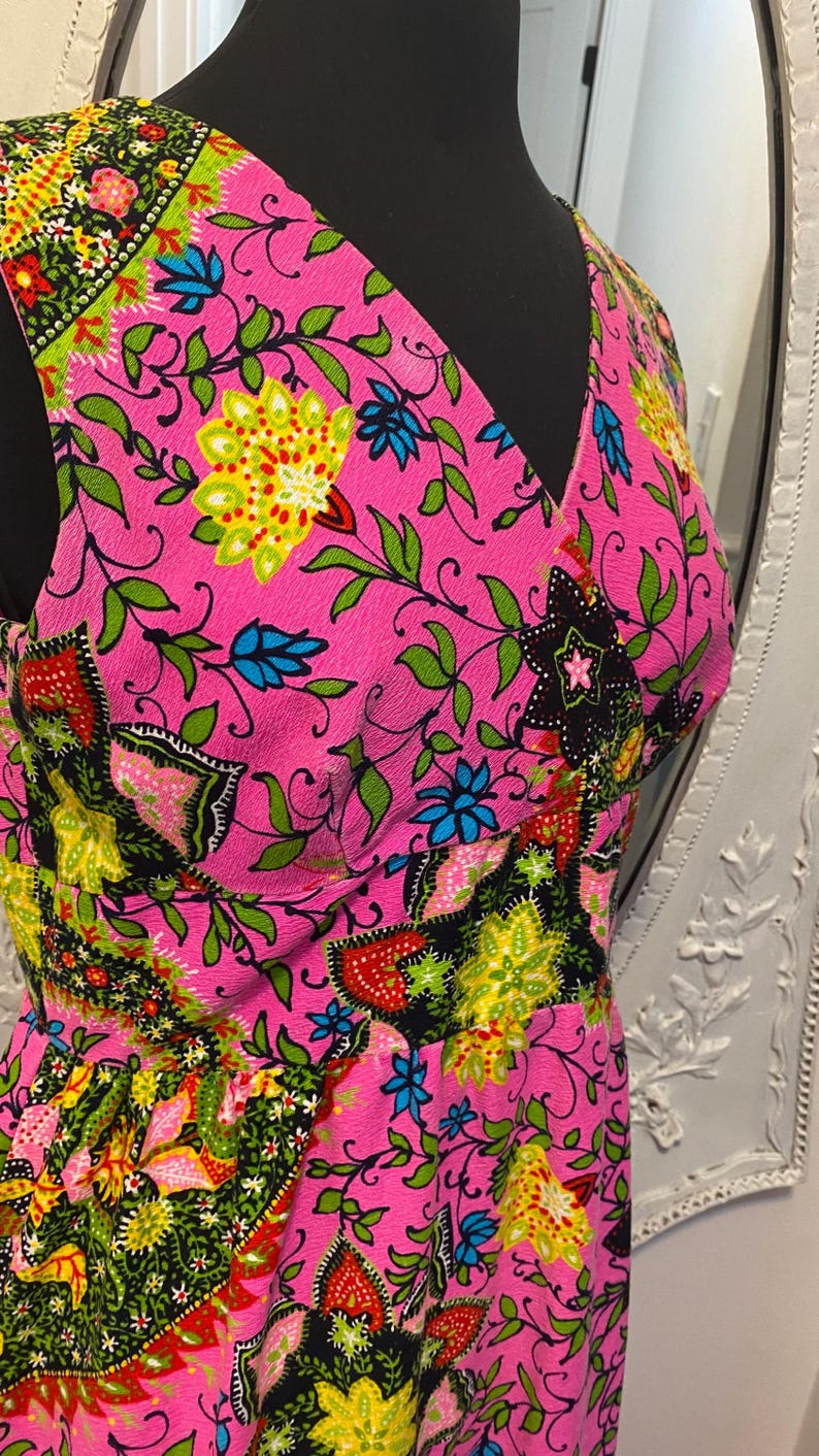 May include: A sleeveless pink dress with a V-neckline and a colorful floral print. The print features green, yellow, blue, red, and black flowers and leaves.