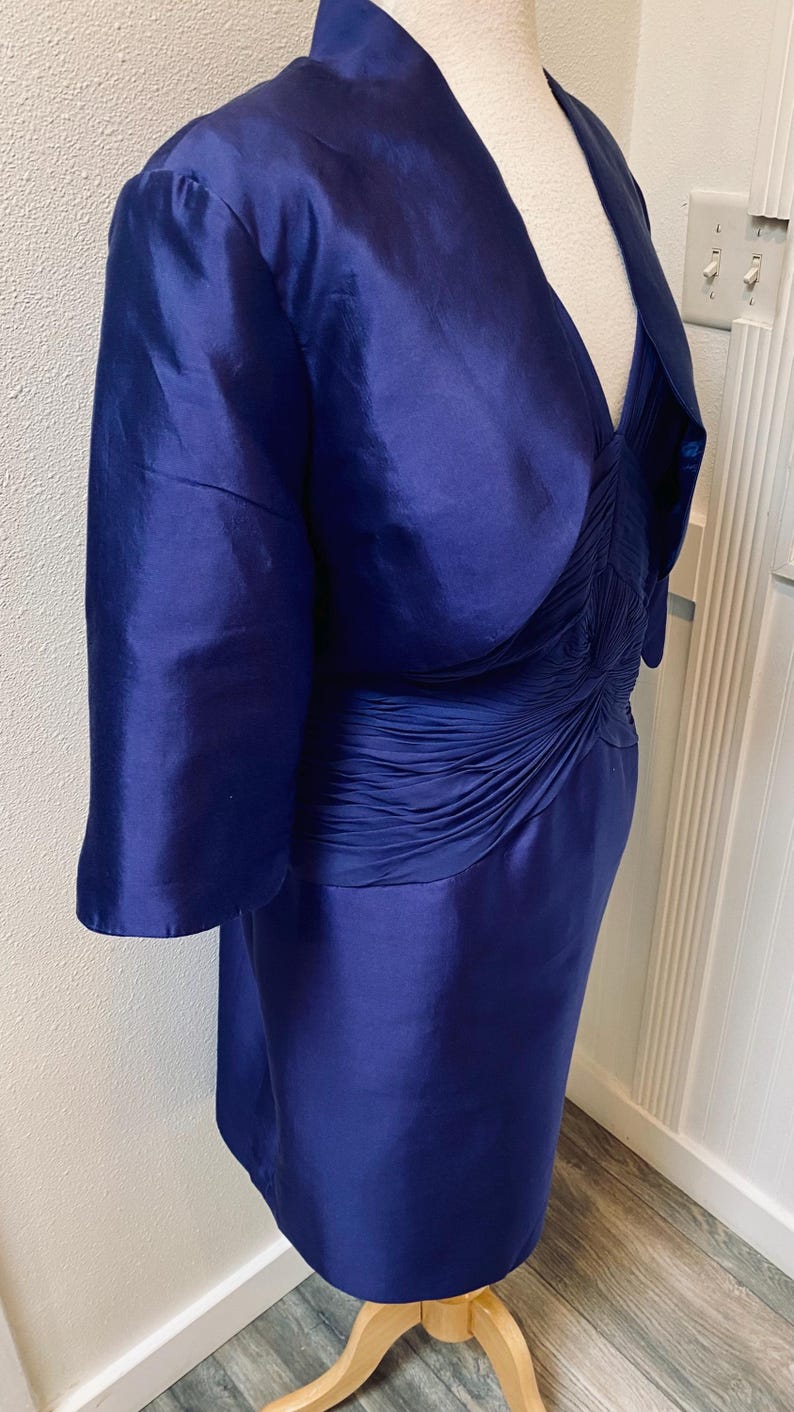 May include: A royal blue dress with a matching jacket. The dress has a fitted bodice with a gathered detail at the waist and a flowing skirt. The jacket has a shawl collar and three-quarter sleeves.
