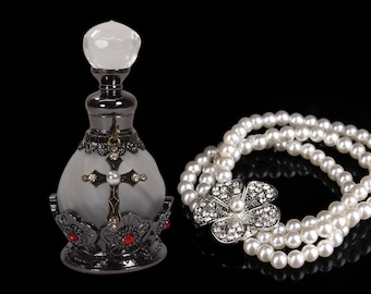Può includere: Un flacone di profumo nero e argento con un design a croce e gioielli rossi. Il flacone è accanto a una collana di perle con un ciondolo a forma di fiore d'argento.