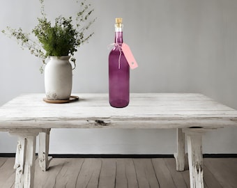 Puede incluir: Una botella de vidrio púrpura con un tapón de corcho y una etiqueta rosa atada alrededor del cuello. La botella está sentada sobre una mesa de madera blanca.