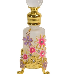 May include: A clear glass bottle with a gold top and a decorative stand. The stand is adorned with pink and purple flowers with gold accents and rhinestones.