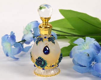 May include: A white frosted glass perfume bottle with gold and blue accents. The bottle has a clear glass stopper with a blue flower design. The bottle is surrounded by blue flowers.