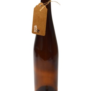 Puede incluir: Una botella de vidrio marr&oacute;n con un tap&oacute;n de corcho y una etiqueta de papel marr&oacute;n atada alrededor del cuello. La etiqueta tiene un dise&ntilde;o floral blanco.
